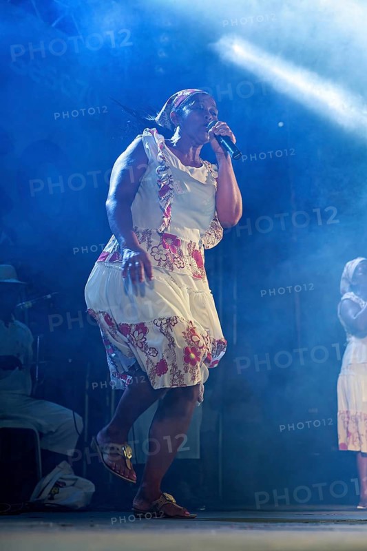 Woman in a floral dress sings impressively on a stage under blue light and smoke, a band plays and dances at a festival in Petrolina in Brazil