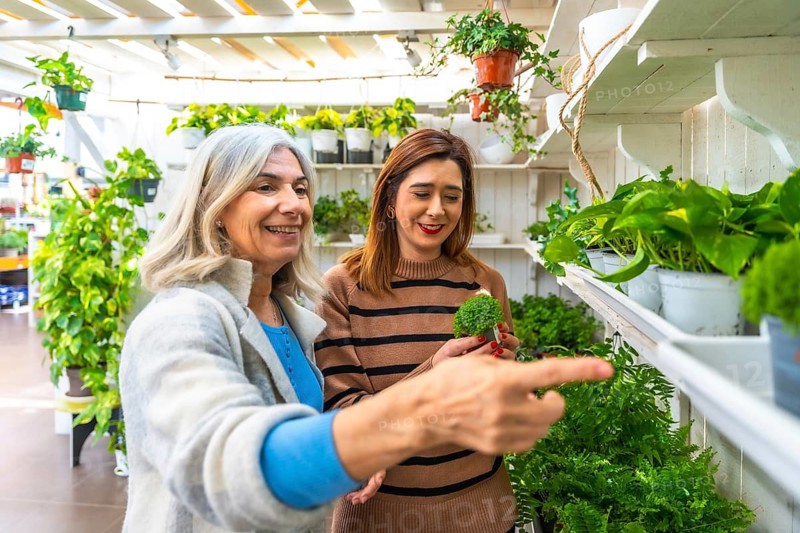 Two smiling women, an elderly mother and her adult daughter, enjoying quality time together in a lush greenhouse, carefully selecting plants for their home or garden
