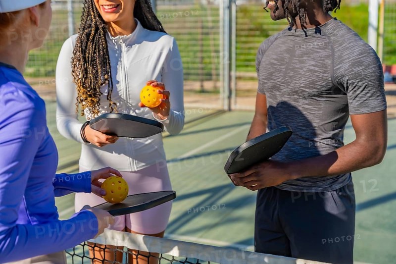 Three diverse adults standing near a net on a pickleball court, holding paddles and balls, engaging in conversation and enjoying an active outdoor sport together on a sunny day
