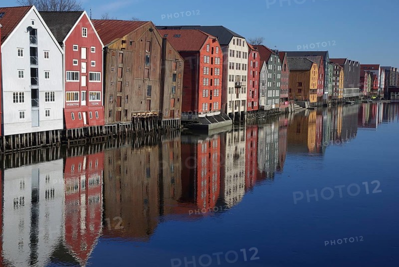 Row of colorful buildings reflected in calm water with clear sky, Trondheim, Nidelva, Tröndelag, Norway