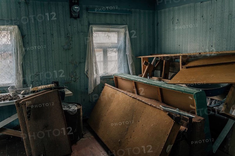 The room of an abandoned old village house with a pile of old broken furniture and other rubbish. An old abandoned room shows worn furniture, peeling walls and scattered objects. Two windows with thin curtains let in soft light, revealing overturned chairs, a damaged bench and stacked wooden boards. The scene conveys decay, disuse and the passage of time in a neglected interior