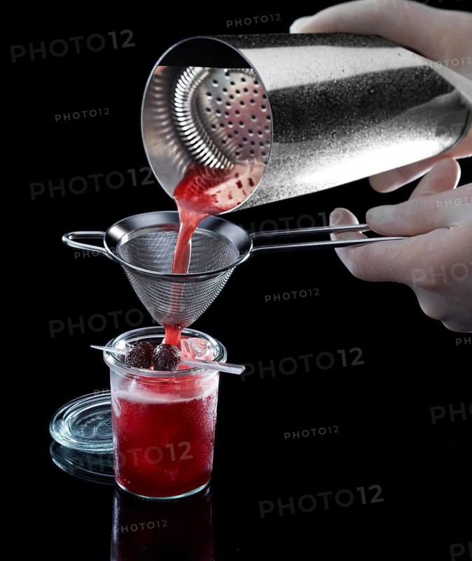 Red cocktail is poured through a sieve, against a black background, 2 berries as garnish, studio shot