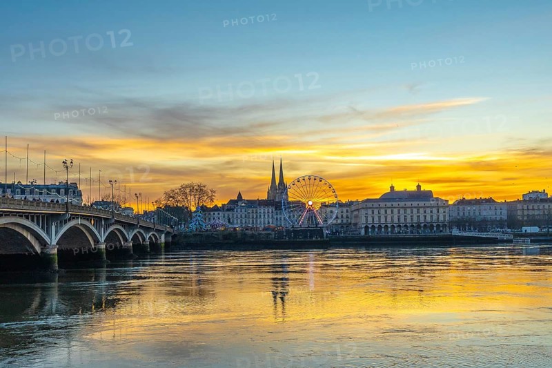 Bayonne cityscape along the adour river at christmas, featuring a illuminated ferris wheel, cathedral spires, and a bridge under a colorful sunset sky reflecting on the water