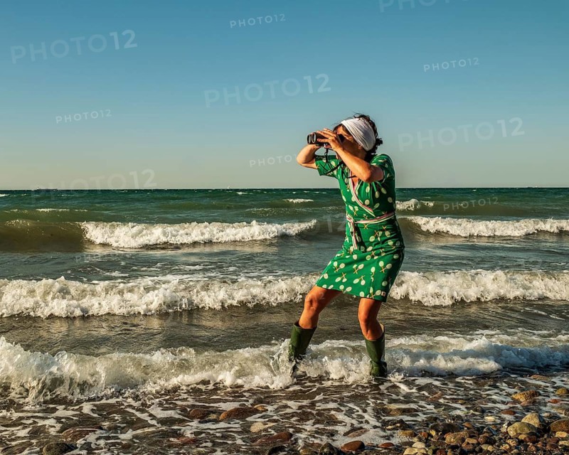 A woman (54) wearing a green tennis dress and green wellington boots is standing in the Baltic Sea with a pair of binoculars, gazing into the distance, Baltic Sea, Germany