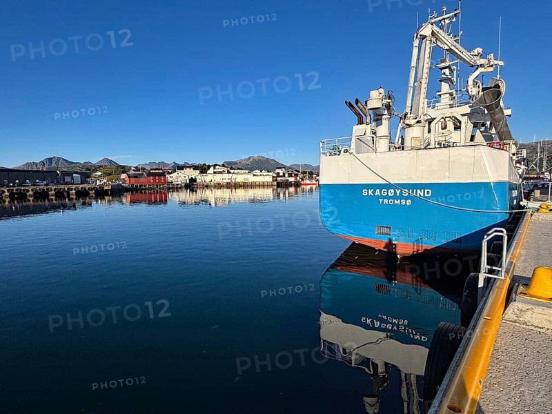 A large ship is lying in the still water of a harbor surrounded by mountains under a clear blue sky, Sommaroy, Sommarøy, Troms, Norway