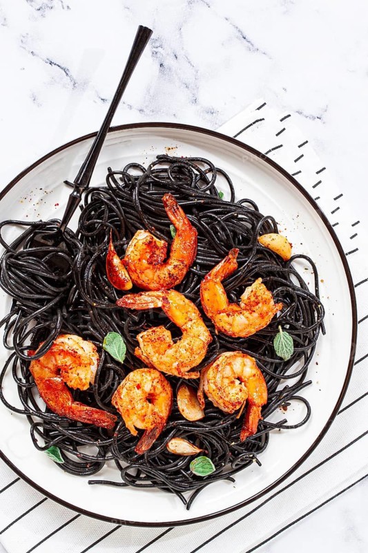 Black spaghetti with shrimp, on a white plate with a fork, on a striped napkin, marble background, homemade, no people