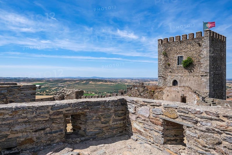 Castle walls with tower and flag offer a wide view of the surrounding countryside under blue skies, Monsaraz, Évora, Portugal