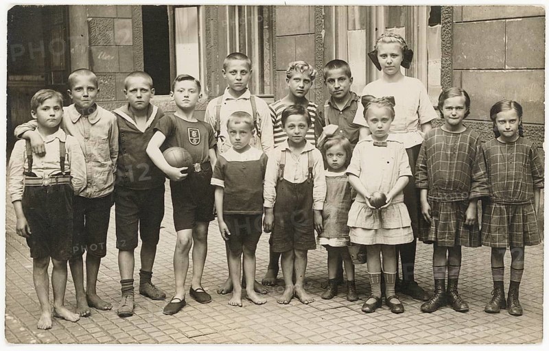 Children, backyard, twenties, big city, poverty, without shoes, barefoot, girls in similar clothes, siblings, group of boys and girls, some barefoot, pose together. Someone is holding a ball. Old-fashioned clothing, around 1920