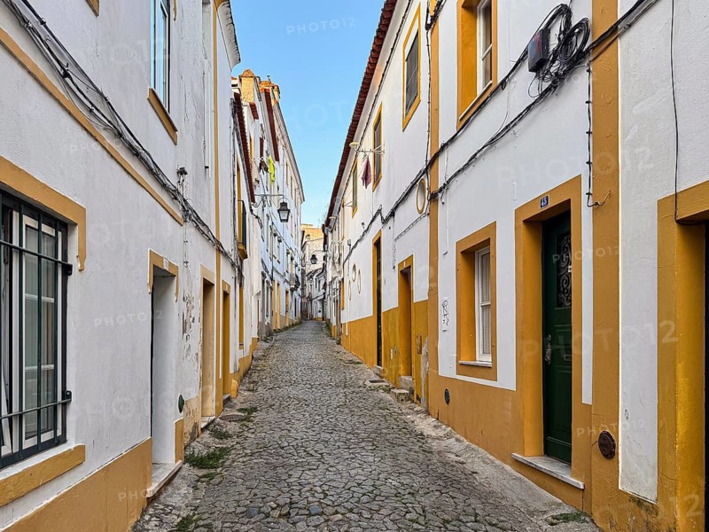Paved alleyway in a narrow alley between white houses with yellow accents in a quiet old town backdrop, Évora, Portugal