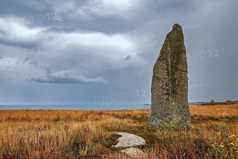 A menhir rises in a meadowy coastal landscape with a view of the sea under dramatic clouds, menhir by Cam Louis, a megalithic monument from the Bronze Age in Plouescat, Brittany