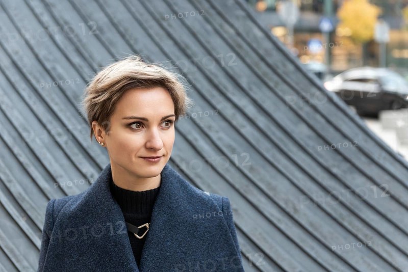 Portrait of an attractive short-haired blonde woman in the city in autumn. A woman with short light brown hair stands outdoors wearing a dark blue coat and black top. Behind her is a large curved architectural structure with ribbed diagonal lines. Blurred urban elements, cars and autumn trees appear in the background. The scene combines a clean portrait with a textured modern city environment