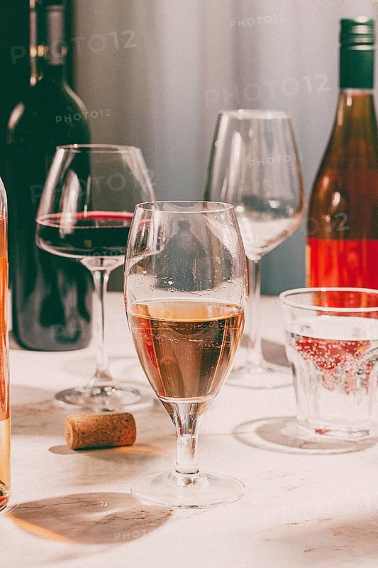 Different types of wine are served in glasses along with wine bottles on a table. A cork is placed nearby. The setting looks inviting and relaxed