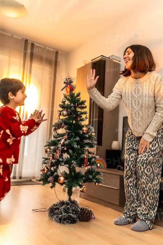 Mother and son in matching pajamas laugh and high five beside a decorated christmas tree in their cozy living room, celebrating holiday joy and warm family bonding at home