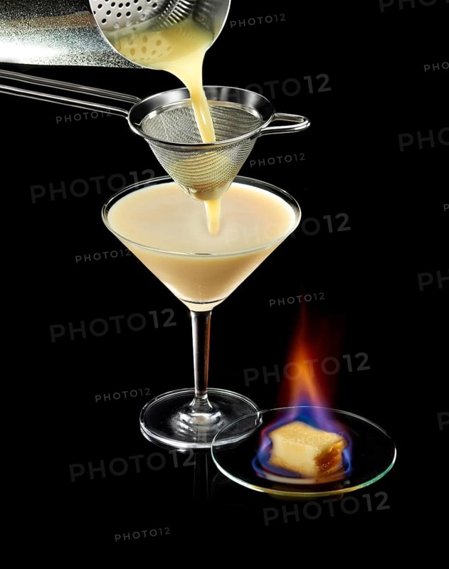 Light brown cocktail is poured through a sieve, against a black background, flambéed confection as decoration, studio shot