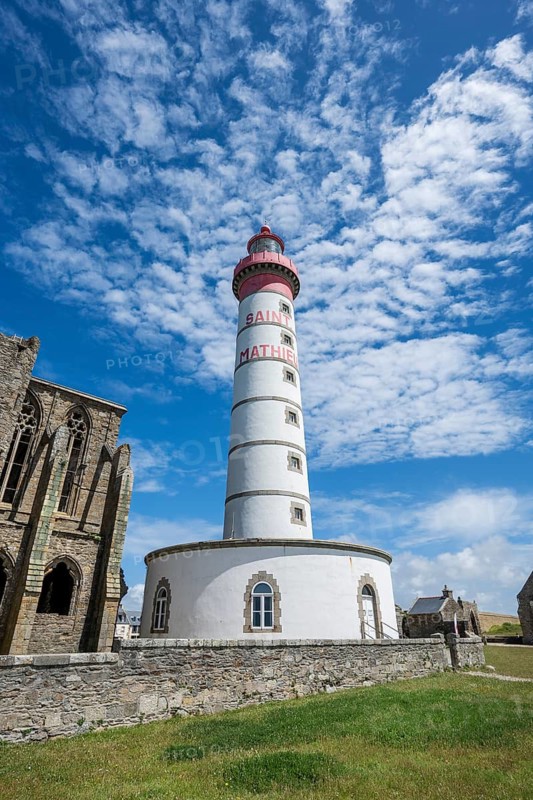 View of the 37 meter high, red-and-white striped lighthouse Phare de Saint-Mathieu, which rises majestically amidst the historic ruins of an old medieval former Benedictine abbey, Abbey Saint-Mathieu de Fine-Terre. A paved footpath leads through a green coastal landscape under blue summer skies with white clouds directly to the historic landmark. Pointe Saint-Mathieu, Plougonvelin, Arrondissement of Brest, Finistère Department, Brittany Region, France