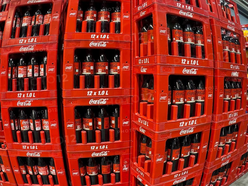 Perspective dynamically deformed photo of left column stacked in boxes 1 liter PET bottles recyclable plastic containers polyethylene terephthalate with caffeinated soft drink Coca-Cola right next to it with black lettering Coca-Cola Zero sugar-free effervescent drink, also known as Coke Zero, Germany