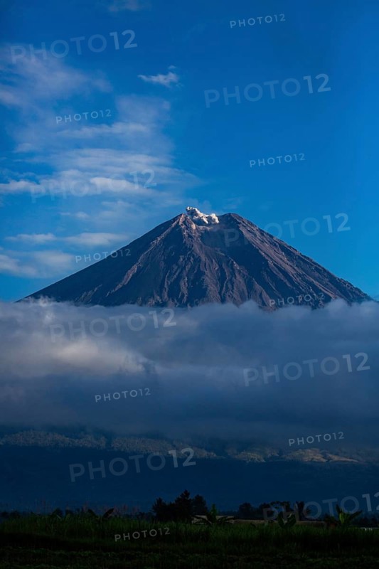 A majestic volcano rises above clouds and a blue sky with light snow cover at the summit, Smoking Volcano, Sumatra, Jakarta, Indonesia