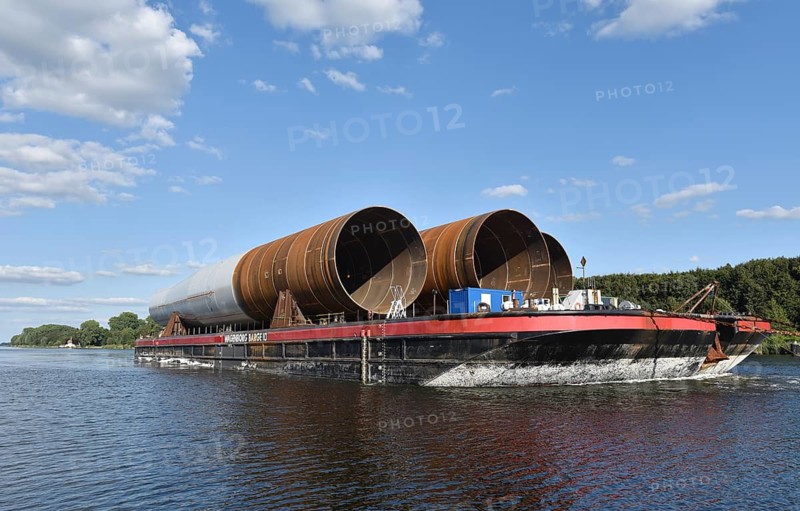 A barge transports large pipes on a river surrounded by trees under a blue sky, tugboat GROSSNORDSEE transports monopiles for wind power foundations in the Kiel Canal