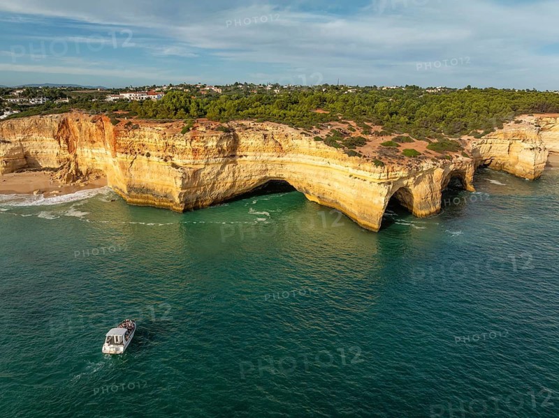 Drone view of Benagil Cave cliffs in Algarve, Portugal, with a tourist tour boat, limestone sea caves, turquoise Atlantic water, and waves