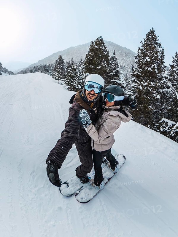 Couple of friends smiling for a selfie while snowboarding down a snowy mountain slope, enjoying an active winter vacation amid pine forests and crisp alpine scenery