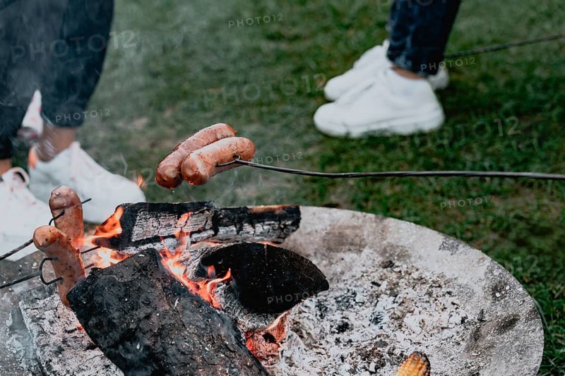 A group of friends roasts sausages on metal skewers on a fire. A close-up view of a campfire shows burning logs and glowing embers while sausages are roasted on metal skewers. A piece of corn rests near the edge of the fire pit, and two people in white sneakers stand on the grass nearby. The scene captures a casual outdoor cooking moment associated with camping, picnics, and social gatherings