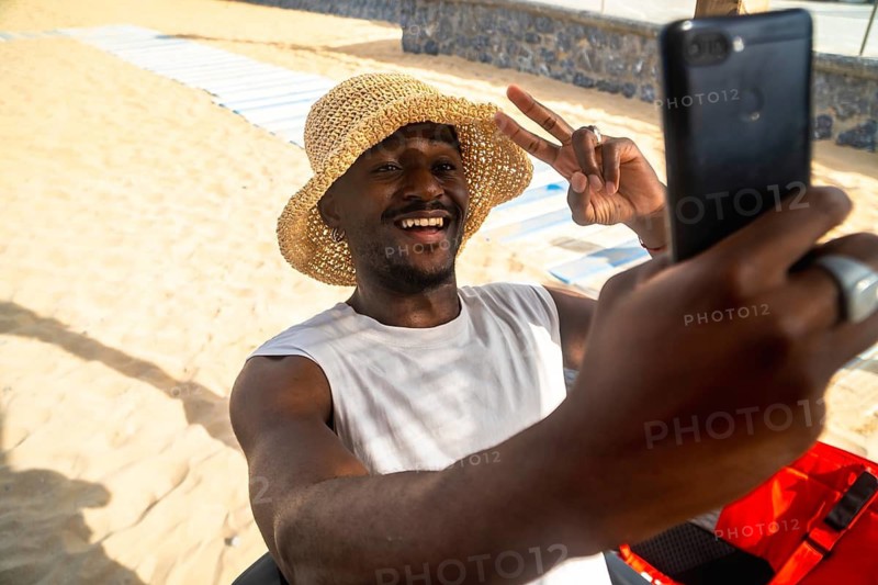 Young black man smiling brightly and making a peace sign while holding a smartphone and taking a selfie on a sunny sandy beach during summer vacation
