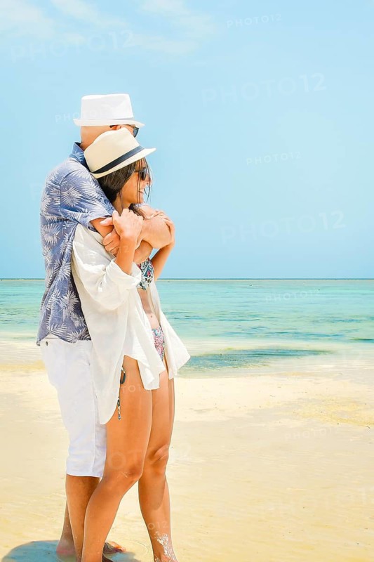 Heterosexual caucasian couple in love husband hugging his wife embracing beach side sand shore turquoise ocean water global tropical island resort destination summer day travel vista
