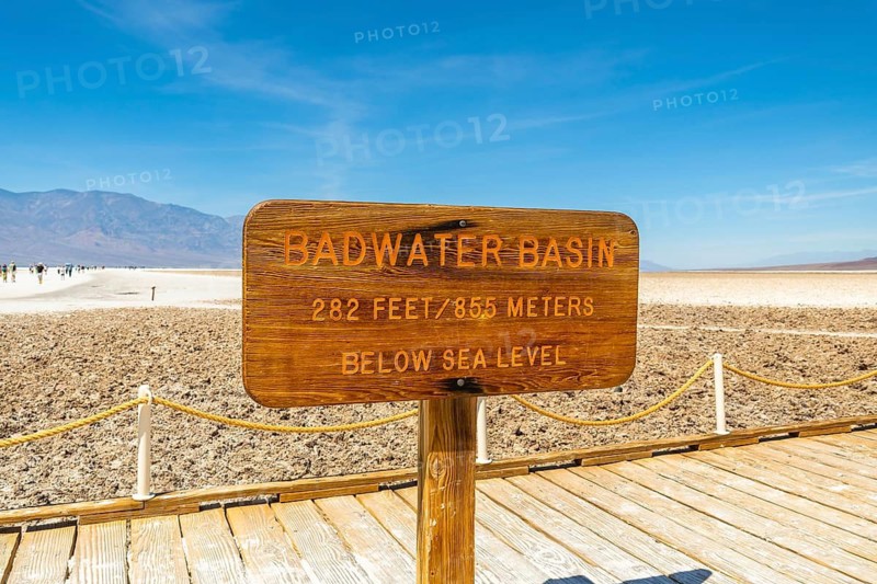 Wooden sign displaying badwater basin, 282 feet - 855 meters - below sea level, situated on a boardwalk in the vast salt flats of death valley national park under a clear blue sky