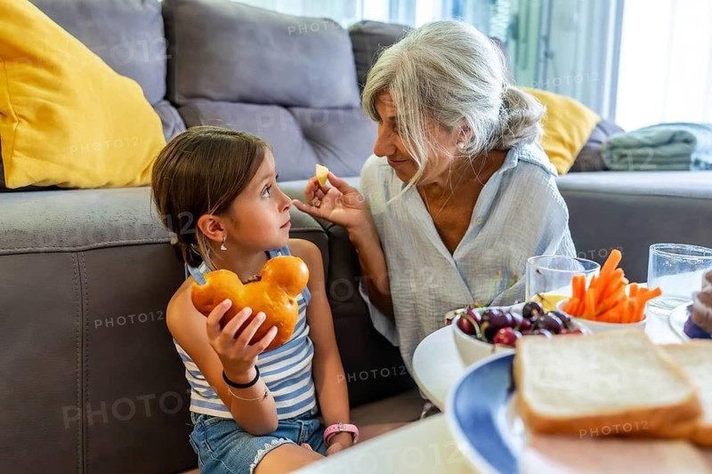Granddaughter and grandmother enjoying an afternoon snack time together, sitting on the floor in a relaxed home environment while sharing healthy food and happy family moments