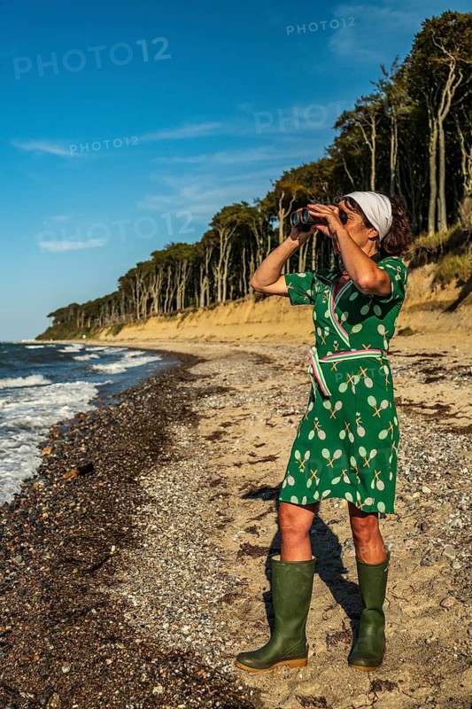 A woman (54) in a green tennis dress and green wellies is standing on the beach with a pair of binoculars, gazing into the distance, Gespensterwald, Nienhagen, Germany