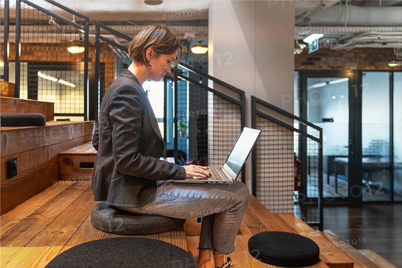 Side view of young woman in jeans and blazer typing on laptop in modern coworking space with wooden steps
