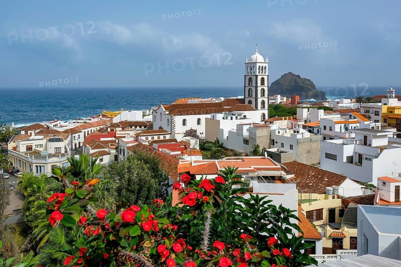 Garachico, Santa Cruz, Tenerife, Canary Islands, Spain - Garachico is a small, tranquil, picturesque town right by the sea in the northwest of the island. City overview with residential buildings, Christ thorn, colorful flowers, palm trees, the Iglesia de Santa Ana church and the Roque de Garachico rock in front