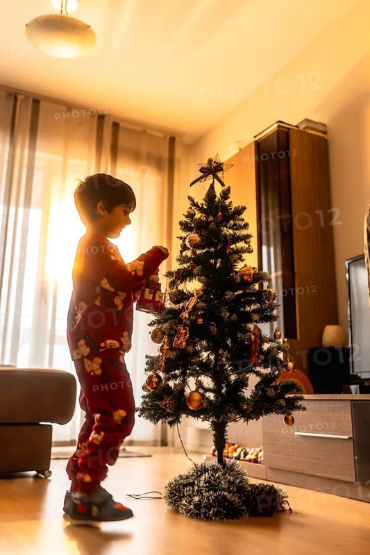 Young boy in pajamas carefully hanging ornaments on a small christmas tree, preparing for joyous celebrations in his cozy home filled with warm holiday light