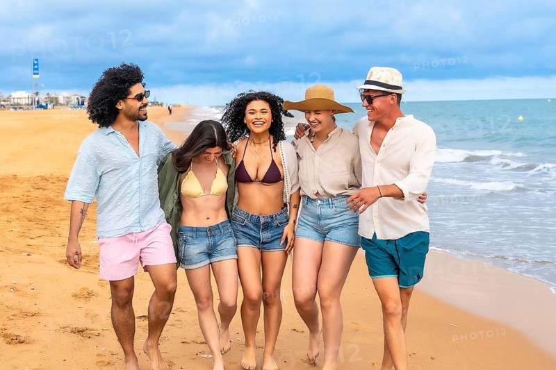 Young multi ethnic friends walking barefoot along a sunny beach, laughing and arm in arm, enjoying a carefree summer day of travel, friendship and seaside relaxation