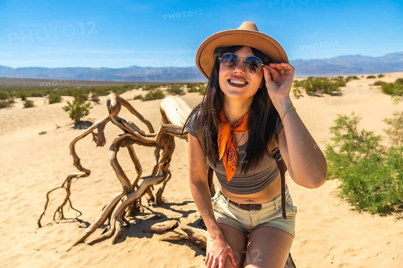 Woman smiling and adjusting sunglasses while sitting on mesquite flat sand dunes in death valley national park under a clear blue sky, enjoying desert adventure and open arid landscape