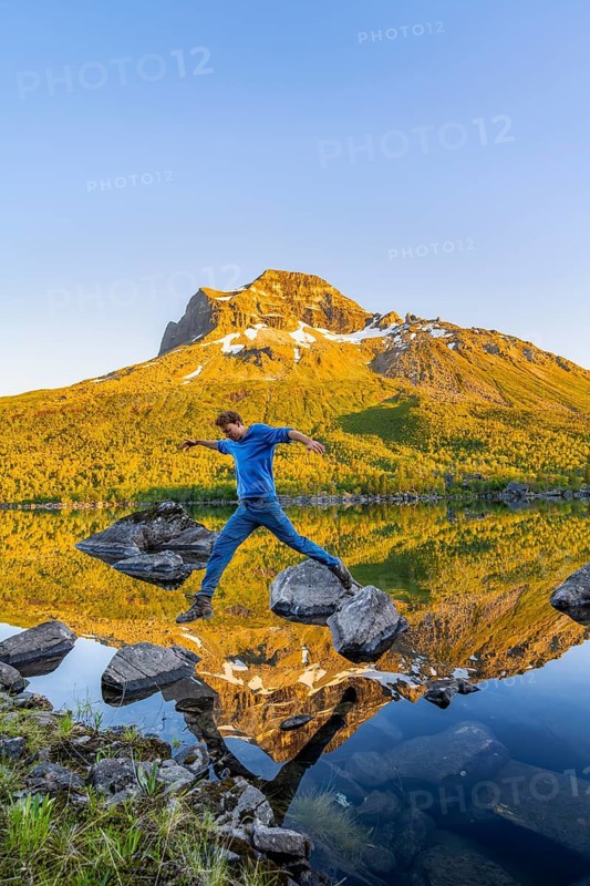 Young man jumping from stone to stone in water, picturesque mountain landscape in the Innerdalen high valley, mountain peaks reflected in Lake Innerdalsvatna, in the evening light at sunset, Trollheimen Mountain Area, Sunndal, Møre og Romsdal, Norway