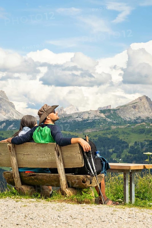 Caucasian couple hikers sit on hand made wooden bench, admiring the majestic Dolomites mountain range in Italy. A peaceful moment of adventure, wellness, and scenic trekking in South Tyrol