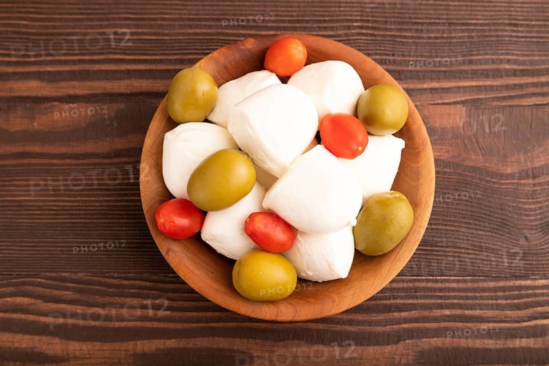 White Mozzarella cheese, with tomatoes and olives in wooden bowl on brown wooden background, top view, flat lay, close up