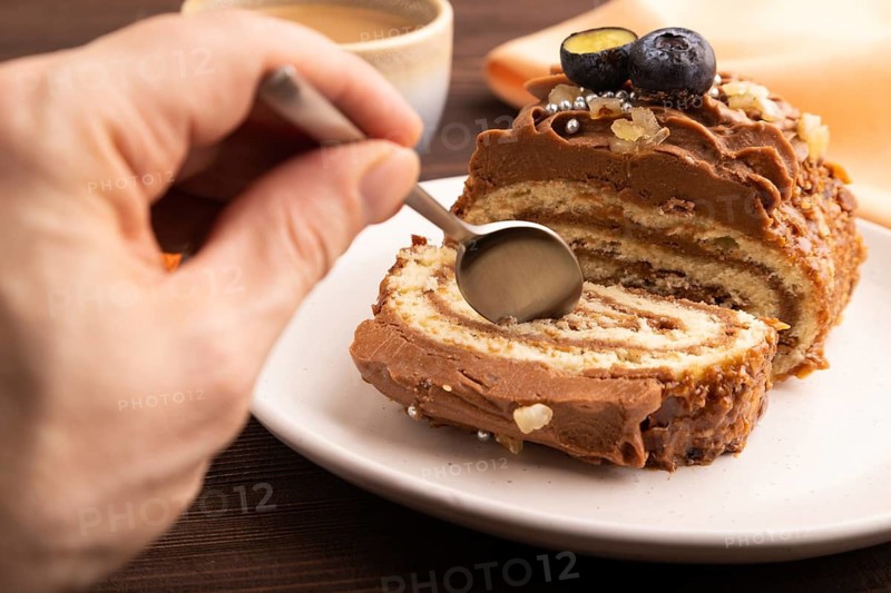 Sweet loaf with cream and caramel with hand, cup of coffee, on brown wooden background with orange textile, close up, side view, selective focus