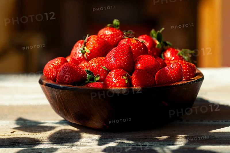 Close-up view of bowl on wooden table with plenty of fresh strawberries inside. Bright daylight shines on fruit, highlighting red color and green leaves. Setting brings focus to bowl