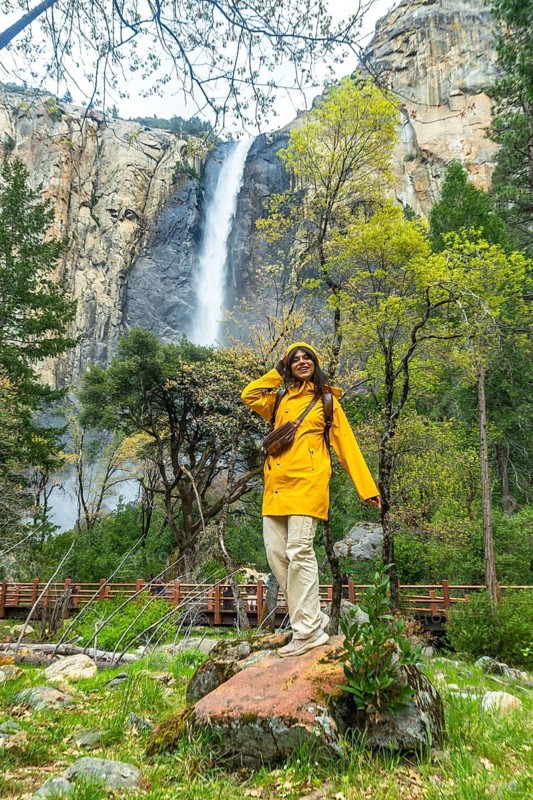 Woman wearing yellow raincoat standing on a rock, admiring the powerful bridalveil fall and lush green forest while hiking in yosemite national park, experiencing nature