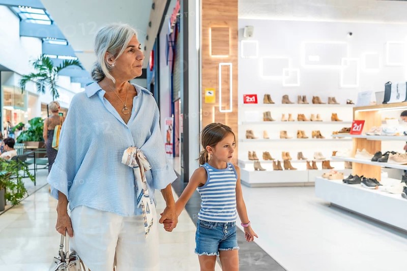 Grandmother and her young granddaughter holding hands and walking through a bright shopping mall, browsing shoe stores and enjoying quality time together