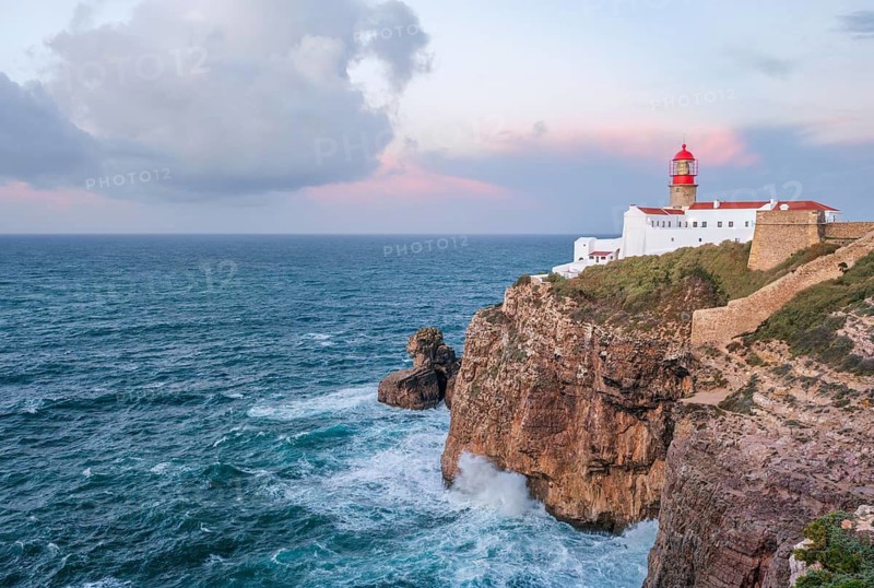 View of the Lighthouse of Cabo de Sao Vicente in Sagres, Algarve, Portugal, at sunrise with Atlantic ocean, cliffs, and orange cloud and cliff