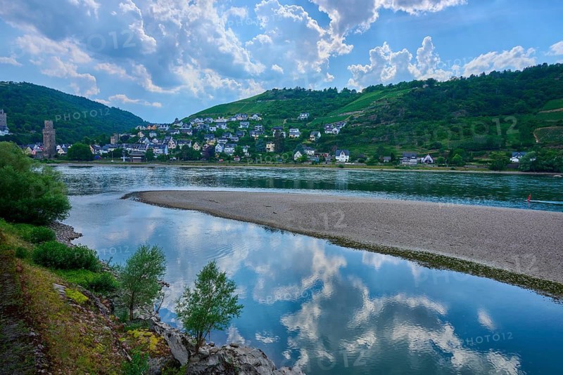A quiet village stretches across hills on the banks of a river with a sandbar, under a cloudy sky, Bacharach am Rhein, Rheingau, Hesse, Germany