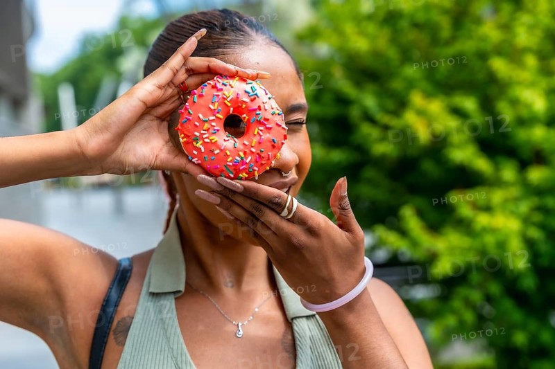 Young woman smiling while holding a vibrant red donut with rainbow sprinkles over one eye, expressing joy and a playful mood outdoors