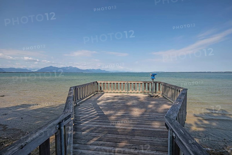 Daytime horizontal view along a wooden observation platform with a blue coin-operated telescope overlooking the clear waters of Chiemsee towards the distant Chiemgau Alps. Chiemgau, Bavaria, Germany