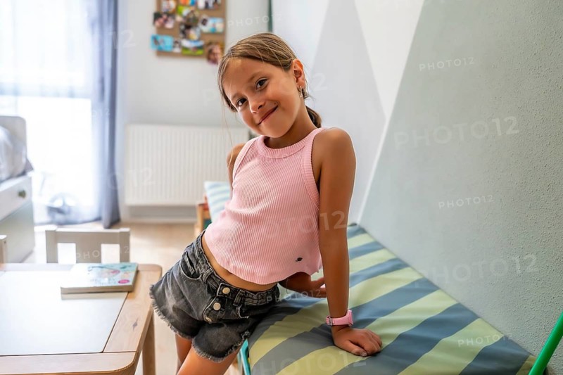 Cheerful young caucasian girl sits smiling on striped bed inside bright modern children's bedroom with wooden furniture