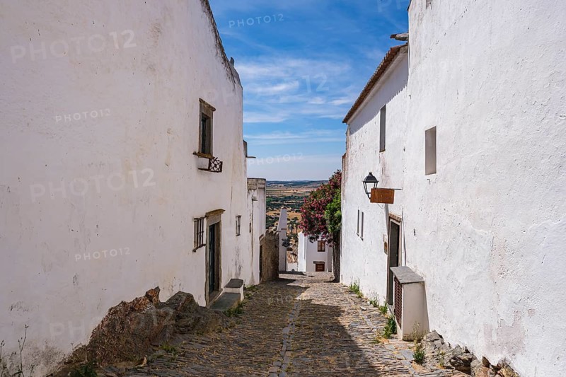 Paved alley between white houses, opens up towards a wide view and blue sky, Monsaraz, Évora, Portugal