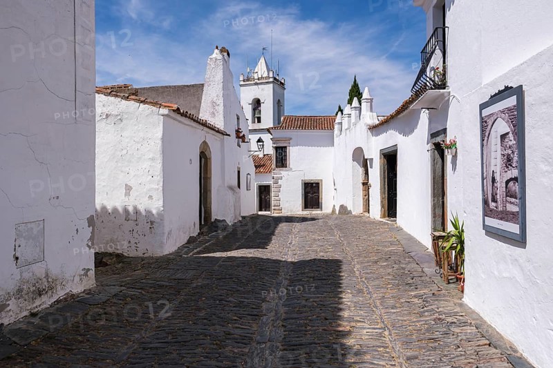 Paved old town street with white buildings and a church, blue sky in the background, Monsaraz, Évora, Portugal