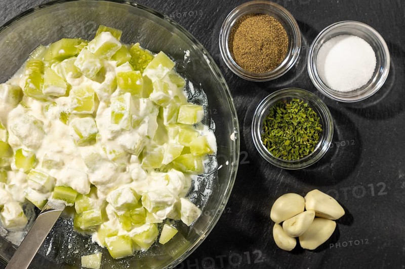 Bowl with sliced cucumber and sour cream, spices and garlic next to it